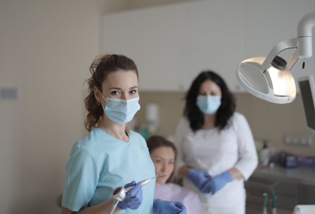 Female dentists in medical masks and latex gloves wearing uniform and treating teeth of patient while standing in modern dental room and looking at camera